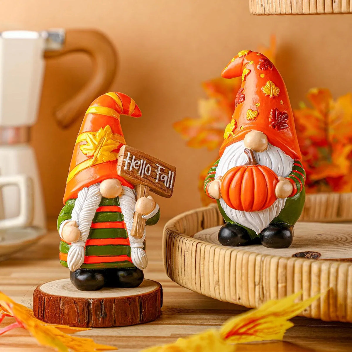 Two decorative autumn gnome figurines styled on a wooden surface with fall leaves and a woven tray. One gnome holds a sign that says 'Hello Fall' and wears a striped green and orange outfit; the other wears an orange pointed hat with leaf patterns and holds a small pumpkin.