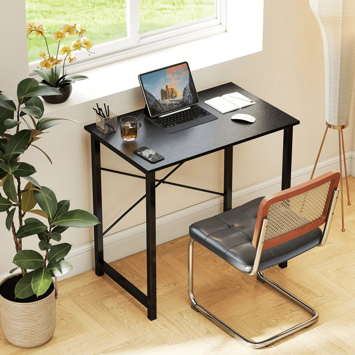 Small black desk with a laptop, mouse, and other items in a room with a window and plant.